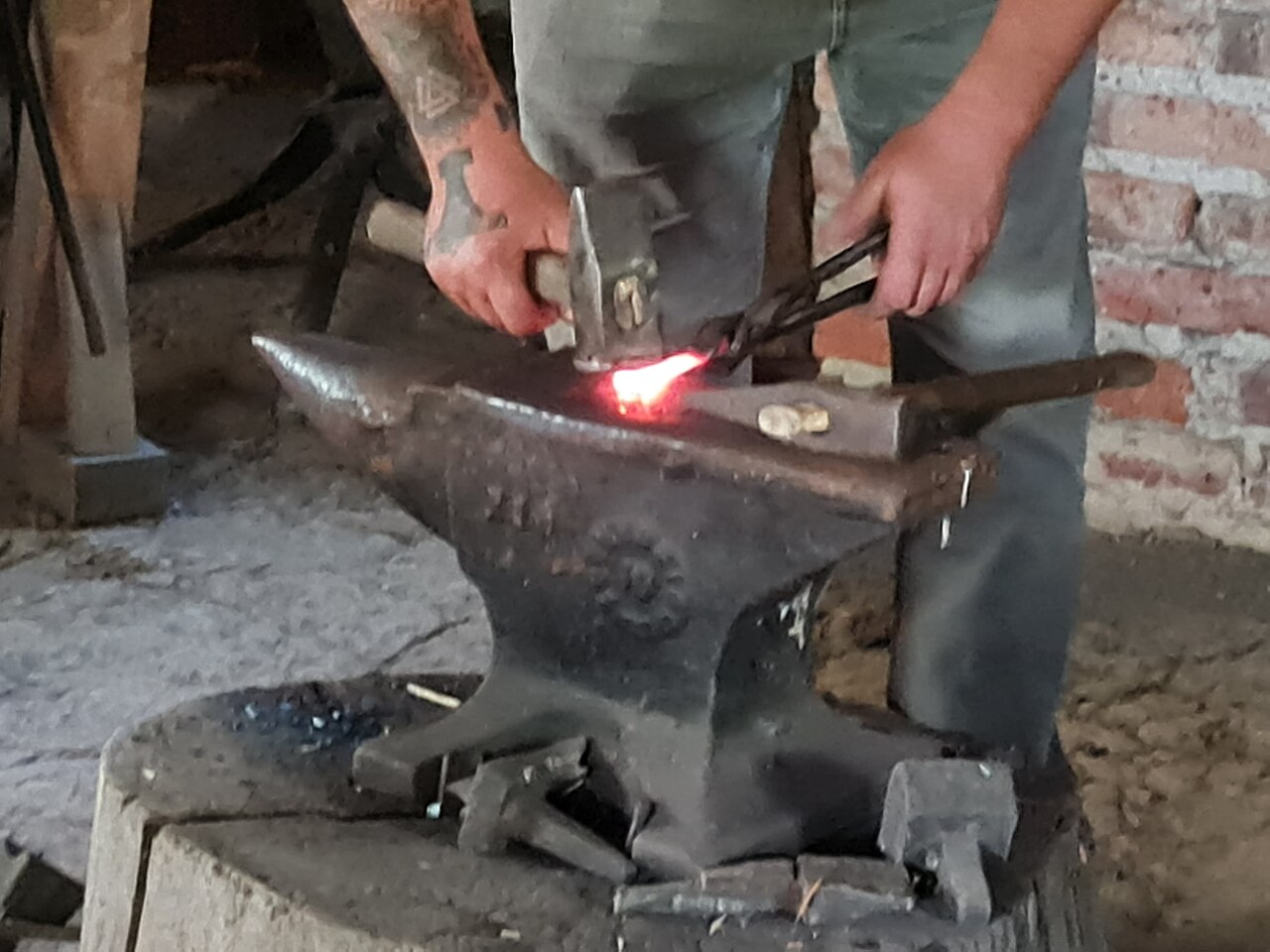 A blacksmith holds a piece of iron glowing orange-red on an anvil with tongs and strikes it with a hammer.